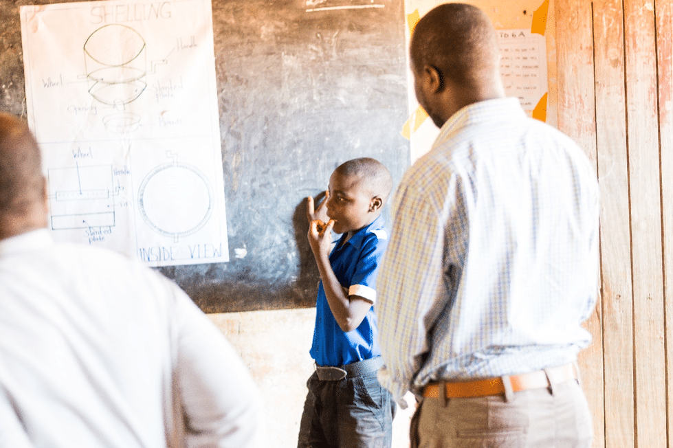 Young boy standing next to blackboard talking with men facing him.